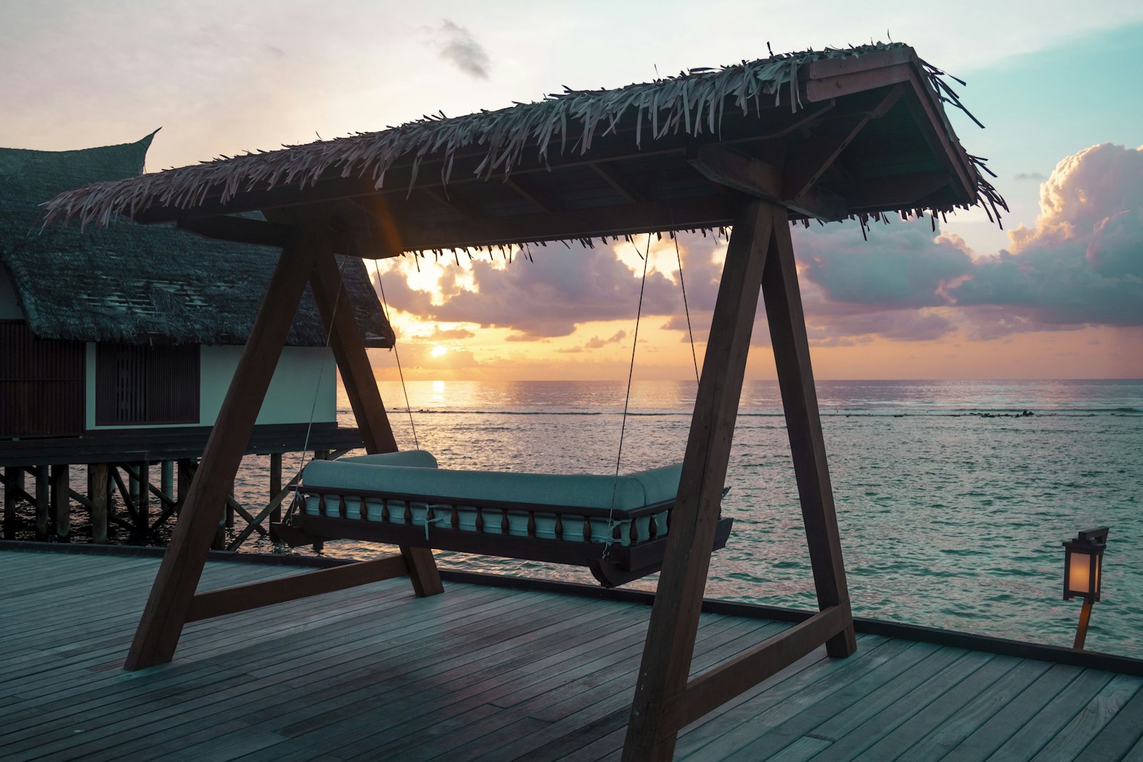 Thatched-roof swing bed on a wooden overwater villa deck at golden sunset over a Maldives lagoon